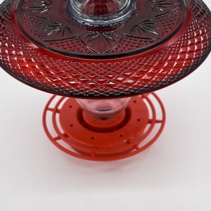 May include: Red glass hummingbird feeder with a decorative, diamond-shaped pattern. The feeder has a red plastic base with a circular perch.