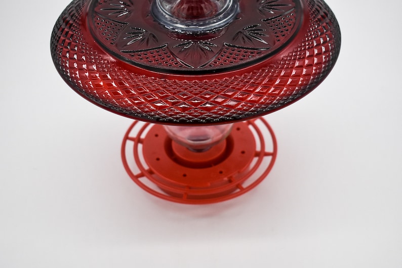 May include: Red glass hummingbird feeder with a decorative, diamond-shaped pattern. The feeder has a red plastic base with a circular perch.