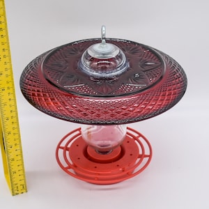 May include: Red glass hummingbird feeder with a clear glass reservoir and a red plastic base. The feeder has a decorative, diamond-shaped pattern.