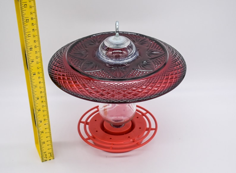 May include: Red glass hummingbird feeder with a clear glass reservoir and a red plastic base. The feeder has a decorative, diamond-shaped pattern.