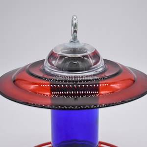 A red and blue glass hummingbird feeder with a clear glass top and a metal hook. The feeder has a red glass base and a cobalt blue glass bottle.