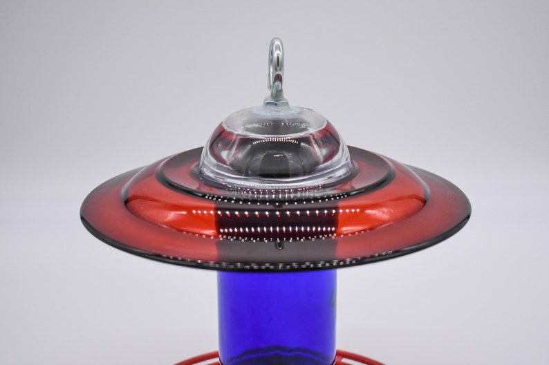 A red and blue glass hummingbird feeder with a clear glass top and a metal hook. The feeder has a red glass base and a cobalt blue glass bottle.
