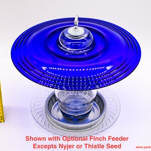 May include: A blue glass bird feeder with a clear glass jar and a metal base. The feeder is shown with an optional finch feeder.  "Shown with Optional Finch Feeder Excepts Nyjer or Thistle Seed" is written below the feeder.