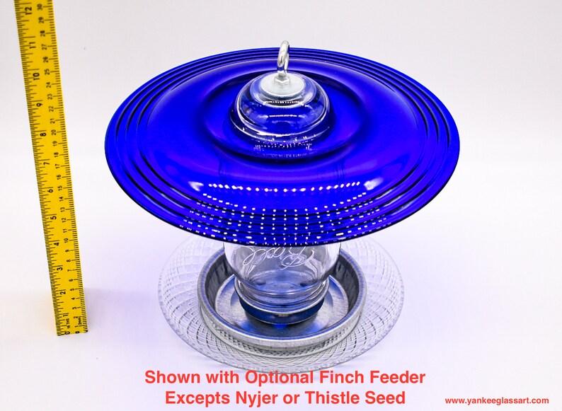 May include: A blue glass bird feeder with a clear glass jar and a metal base. The feeder is shown with an optional finch feeder.  "Shown with Optional Finch Feeder Excepts Nyjer or Thistle Seed" is written below the feeder.
