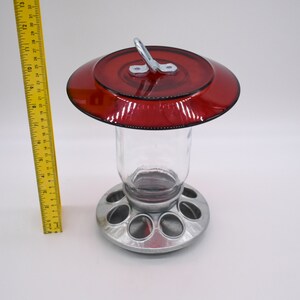 May include: A red glass bird feeder with a metal base and a hanging hook. The feeder has a clear glass jar and a metal base with multiple feeding ports.