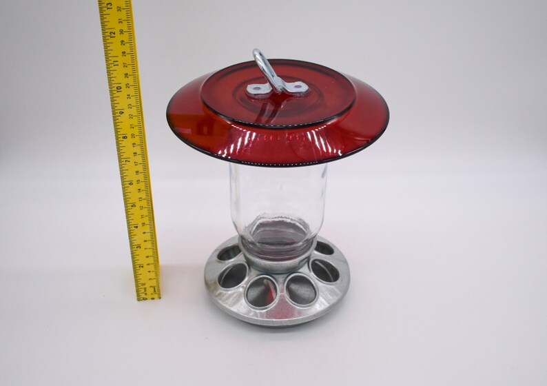 May include: A red glass bird feeder with a metal base and a hanging hook. The feeder has a clear glass jar and a metal base with multiple feeding ports.