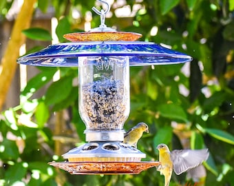 May include: A hanging bird feeder with a blue glass top and a clear glass jar filled with birdseed. The feeder has a decorative orange glass dish at the bottom. Two small yellow birds are perched on the feeder.