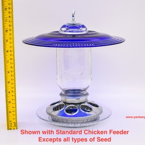 May include: A blue glass bird feeder with a clear glass jar and a metal base. The feeder is shown with a standard chicken feeder.  Shown with Standard Chicken Feeder Excepts all types of Seed www.yankeeglassart.com