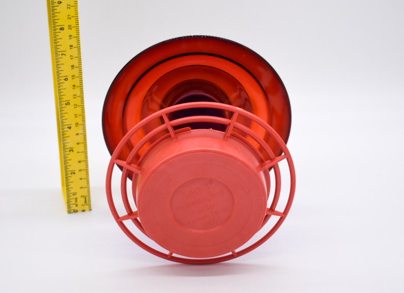 Red hummingbird feeder with a red base and a black and red top. The feeder has a wire cage around the base and the words 'Hummingbird Feeder' are printed on the base.