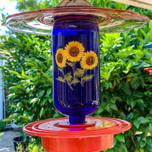May include: A blue glass hummingbird feeder with a red plastic base and a pink glass top. The feeder features a sunflower design.