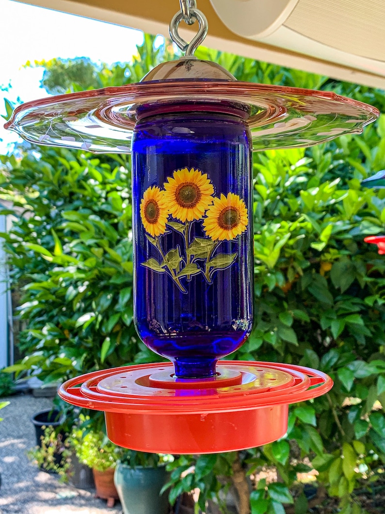 May include: A blue glass hummingbird feeder with a red plastic base and a pink glass top. The feeder features a sunflower design.