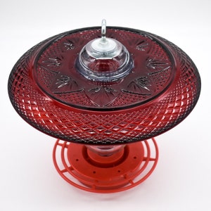 May include: Red glass hummingbird feeder with a decorative, diamond-shaped pattern. The feeder has a clear plastic top with a metal hook for hanging.