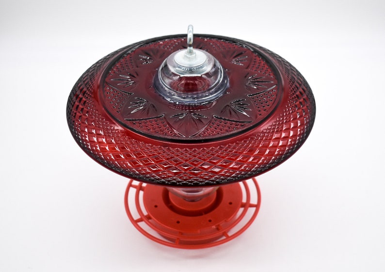May include: Red glass hummingbird feeder with a decorative, diamond-shaped pattern. The feeder has a clear plastic top with a metal hook for hanging.
