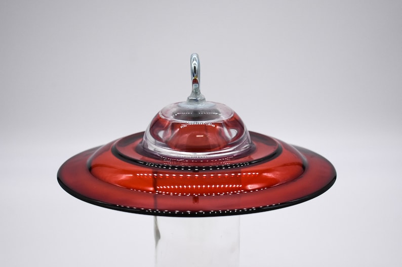 A red glass hummingbird feeder with a clear glass dome and a metal hook. The top of the feeder has a wide, flat base and a narrow, cylindrical neck.