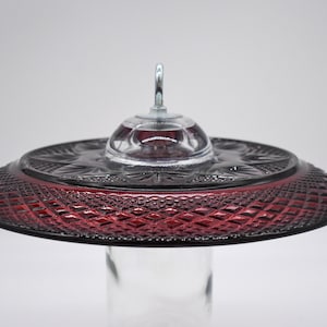May include: A red glass hummingbird feeder with a clear glass dome and a metal hook. The feeder has a diamond pattern on the bottom.