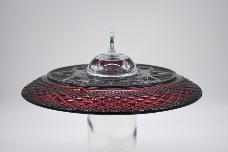 May include: A red glass hummingbird feeder with a clear glass dome and a metal hook. The feeder has a diamond pattern on the bottom.
