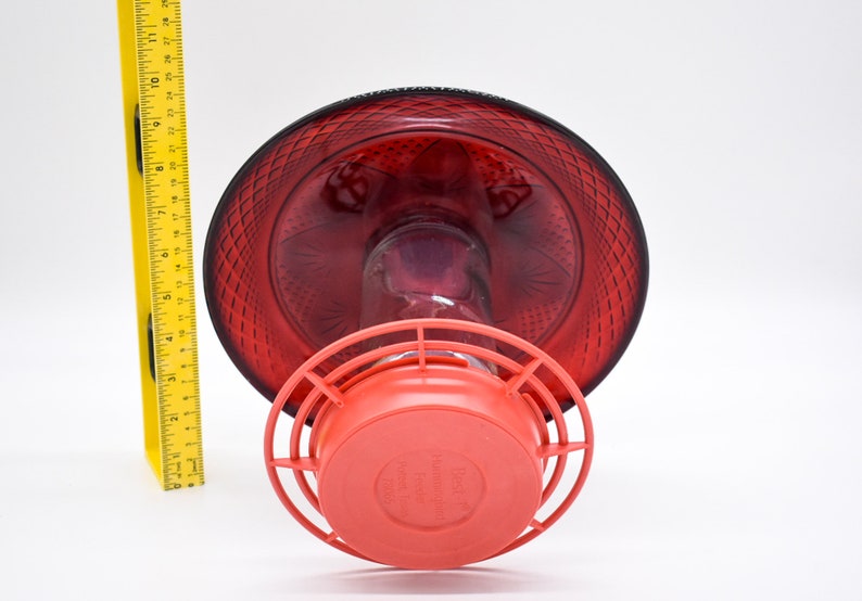 May include: Red glass hummingbird feeder with a red plastic base and a wire cage. The feeder has a decorative diamond pattern.