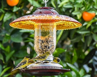 May include: A glass bird feeder with a yellow glass top and a clear glass jar filled with birdseed. Three yellow birds are perched on the feeder. The feeder is hanging from a tree branch.