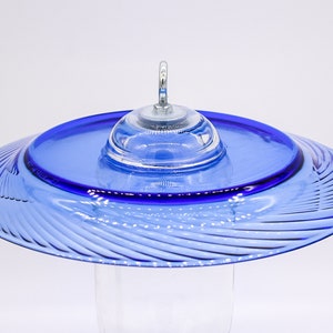 May include: A blue glass hummingbird feeder with a swirling pattern and a clear glass dome on top. The feeder has a metal hook for hanging.