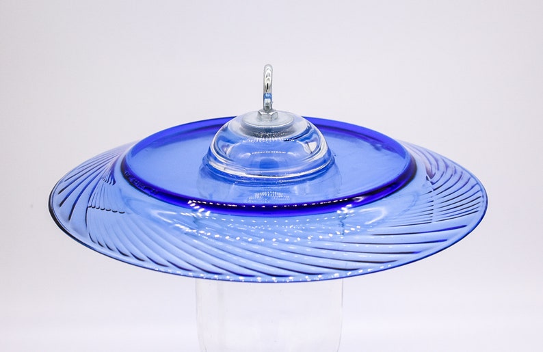 May include: A blue glass hummingbird feeder with a swirling pattern and a clear glass dome on top. The feeder has a metal hook for hanging.