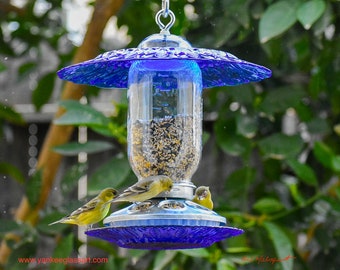 May include: A blue glass bird feeder with a clear glass jar filled with birdseed. Four yellow birds are perched on the feeder, eating the seeds. The feeder is hanging from a metal hook.