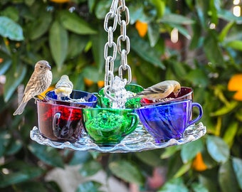 May include: A bird feeder made of colorful teacups hanging from a chain. The feeder has four cups, one red, one green, one blue, and one clear. There are three birds eating from the feeder.