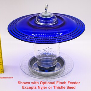 May include: A blue glass bird feeder with a clear glass jar and a metal base. The feeder is designed to hold a mason jar. The feeder is shown with an optional finch feeder.  Shown with Optional Finch Feeder Excepts Nyjer or Thistle Seed