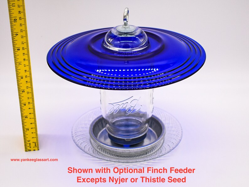 May include: A blue glass bird feeder with a clear glass jar and a metal base. The feeder is designed to hold a mason jar. The feeder is shown with an optional finch feeder.  Shown with Optional Finch Feeder Excepts Nyjer or Thistle Seed