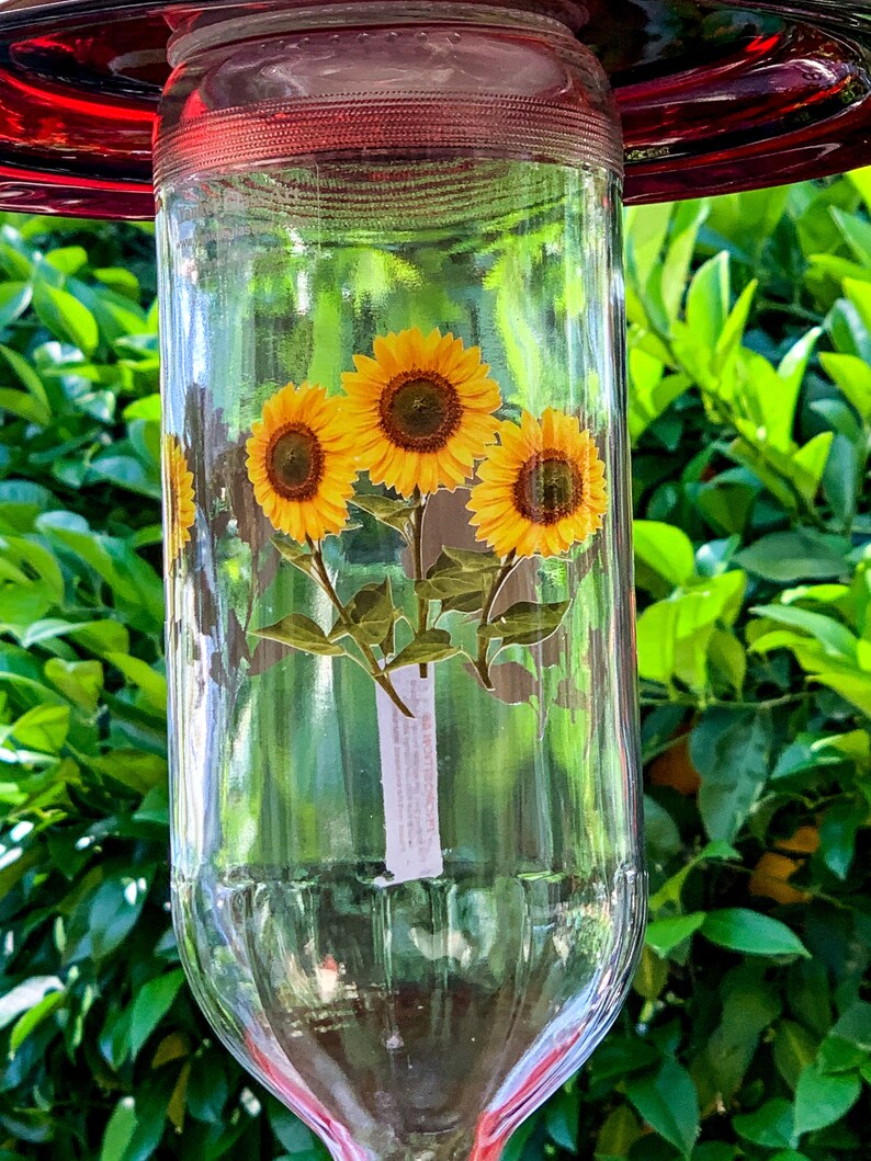 May include: A clear glass hummingbird feeder with a red top and a sticker of three yellow sunflowers on the front.
