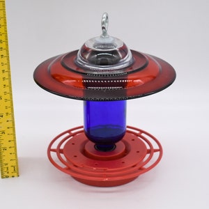Red and blue hummingbird feeder with a clear glass top and a blue glass bottle. The feeder has a red base with a ring of holes around the bottle.