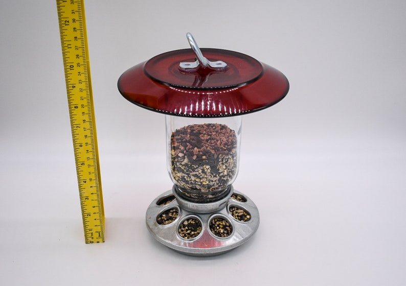 May include: A red bird feeder with a clear glass container filled with birdseed. The feeder has a metal base with multiple feeding ports and a red plastic lid with a metal hook.
