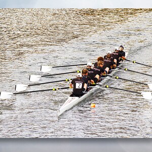 May include: A rowing team of eight rowers in black shirts and black shorts are rowing a white racing shell on a body of water. The rowers are all facing forward and the oars are in the water. The water is rippling and there is a cloudy sky in the background.