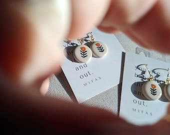 Könnte beinhalten: Ein Paar weiße Keramikohrringe mit einem kleinen orangefarbenen Blumendesign und goldenen Haken. Die Ohrringe befinden sich auf einer weißen Karte mit dem Text "You are beautiful inside and out. MIFAS".