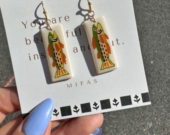 May include: A pair of rectangular earrings with a cream background, featuring a colorful fish design in orange, green, and yellow. The earrings hang from gold-colored hooks. The text "You are beautiful and..." is visible.