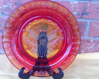May include: A red glass ashtray with a raised design of a bird with outstretched wings and a starburst pattern. The ashtray has a gold-colored rim and a floral design around the edge.