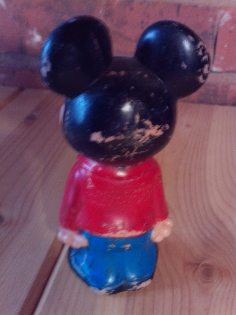 May include: A black and red plastic Mickey Mouse figurine with large ears and blue pants. The figurine has a worn and faded appearance.