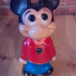 May include: A vintage plastic Mickey Mouse figurine with black ears, a red shirt, and blue pants. The figurine has a friendly expression with large eyes and a wide smile.