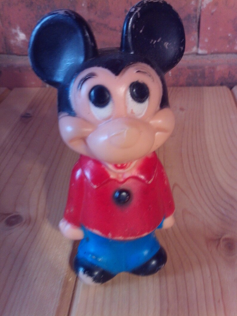 May include: A vintage plastic Mickey Mouse figurine with black ears, a red shirt, and blue pants. The figurine has a friendly expression with large eyes and a wide smile.