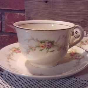 Pode incluir: Um conjunto vintage de chávena e pires com um design floral. A chávena e o pires são brancos com uma borda dourada e apresentam flores cor-de-rosa e amarelas.