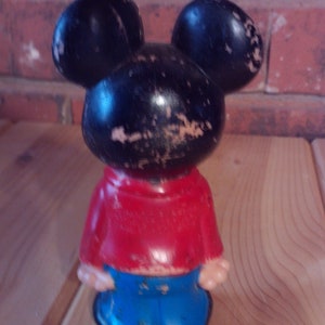 May include: A vintage Mickey Mouse figurine with black ears, a black head, a red shirt, and blue pants. The figurine is made of plastic and has a worn appearance.
