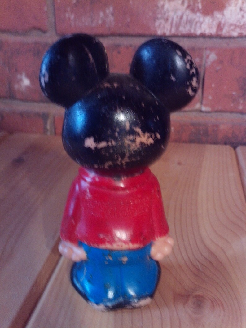 May include: A vintage Mickey Mouse figurine with black ears, a black head, a red shirt, and blue pants. The figurine is made of plastic and has a worn appearance.