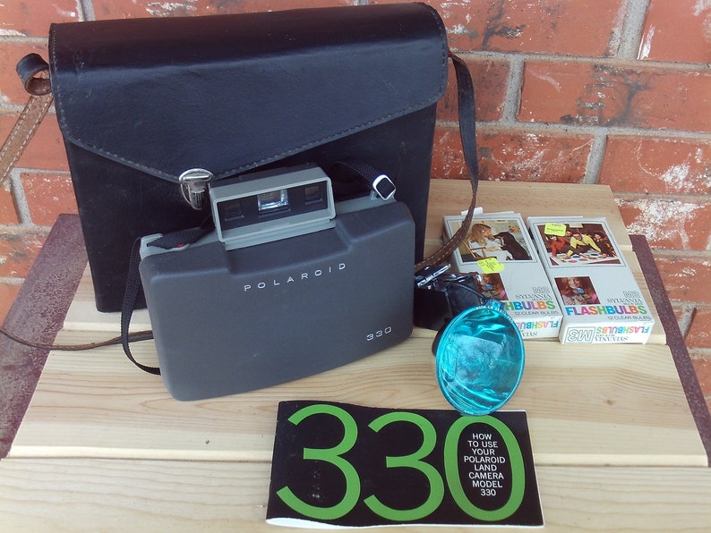 May include: A black leather camera case with a Polaroid Land Camera Model 330 inside. The camera is black and gray with a silver flash attachment. There are two boxes of flashbulbs and a blue flash reflector on the table.