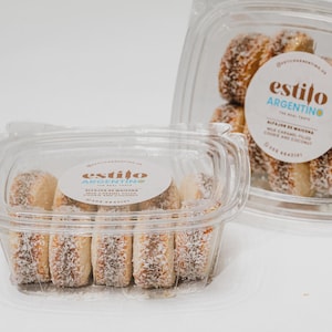 May include: Two clear plastic containers filled with round alfajores de maicena cookies. Each cookie features a caramel filling and shredded coconut edges. The circular white labels on the lids display the text Estilo Argentino, The Real Taste, Alfajor de Maicena, Milk Caramel Filled Cookie and Coconut.