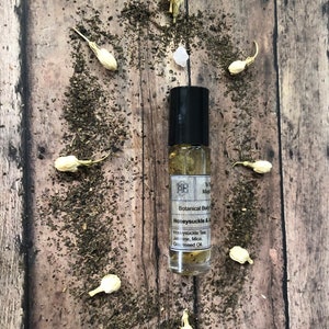 May include: A small glass bottle with a black rollerball top and a white label. The label reads "MB Botanical Body Oil Honeysuckle & Jasmine, Mica, Grapeseed Oil."