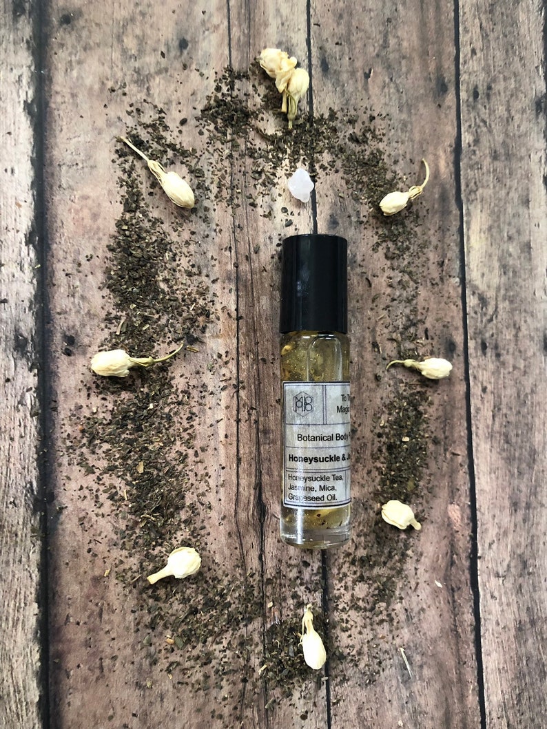 May include: A small glass bottle with a black rollerball top and a white label. The label reads "MB Botanical Body Oil Honeysuckle & Jasmine, Mica, Grapeseed Oil."