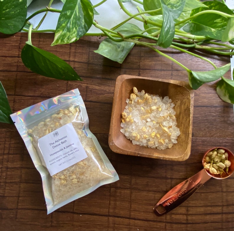 May include: A clear bag and a small wooden bowl filled with bath salts and dried flowers. The bag is labelled "The Alchemist Detox Bath Honeysuckle & Jasmine." A copper measuring spoon and green leafy vines are also present.