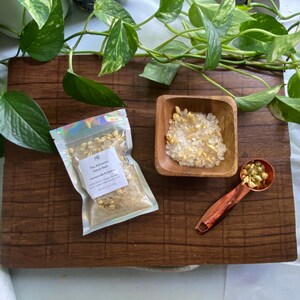 May include: A wooden board displays a small wooden bowl, a copper measuring spoon, and a sealed bag of bath salts. The bag is labelled "The Alchemist Botanic Bath Honeysuckle & Jasmine". Green leaves are arranged around the items.