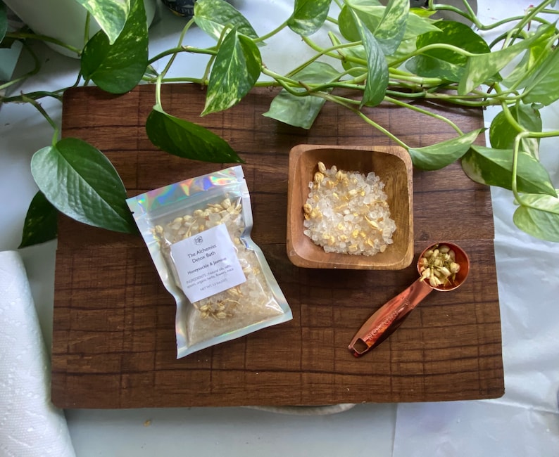 May include: A wooden board displays a small wooden bowl, a copper measuring spoon, and a sealed bag of bath salts. The bag is labelled "The Alchemist Botanic Bath Honeysuckle & Jasmine". Green leaves are arranged around the items.
