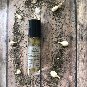 May include: A clear glass roll-on bottle with a black cap and a white label that reads "To The Magic Box Herbal Body Potion Honeysuckle & Jasmine Rose Quartz". The bottle is sitting on a wooden surface with white flower petals and green tea leaves scattered around it.