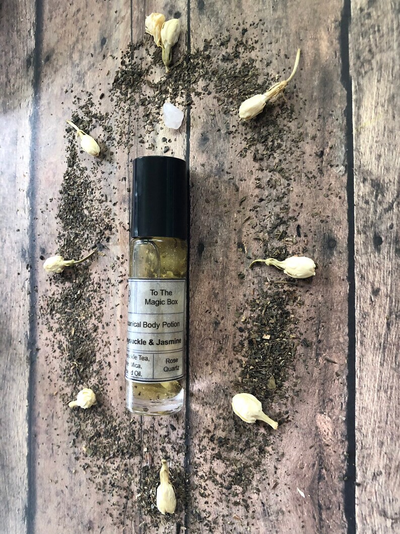 May include: A clear glass roll-on bottle with a black cap and a white label that reads "To The Magic Box Herbal Body Potion Honeysuckle & Jasmine Rose Quartz". The bottle is sitting on a wooden surface with white flower petals and green tea leaves scattered around it.